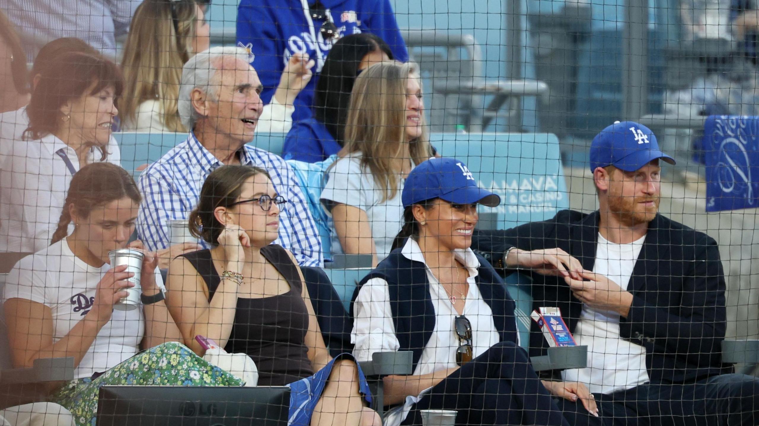 Prince Harry and his wife Megham Markle, wearing Los Angeles Dodgers caps, watch the game sitting in front of Dodgers legend Sandy Koufax